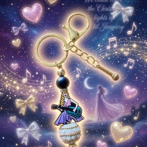 May include: A gold-toned keychain with a charm featuring a woman playing a guitar, a microphone, and a black bead. The charm is adorned with a white and opalescent bead. The background has musical notes, hearts, bows, and text that reads "We could leave the Christmas lights up til January."
