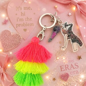 May include: A keychain with a star-shaped charm, a microphone charm, and a figure charm. The keychain also has a three-tiered tassel in neon pink, yellow, and green. The background includes a pink heart with the text "it's me, hi I'm the problem".