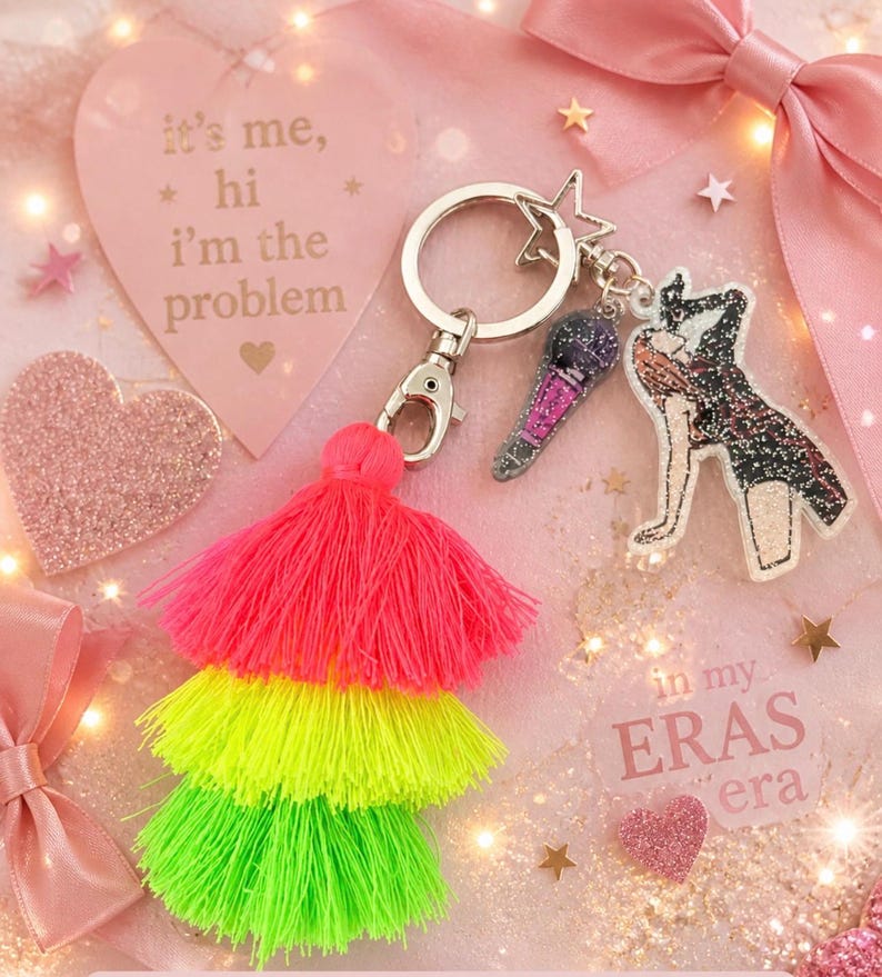May include: A keychain with a star-shaped charm, a microphone charm, and a figure charm. The keychain also has a three-tiered tassel in neon pink, yellow, and green. The background includes a pink heart with the text "it's me, hi I'm the problem".