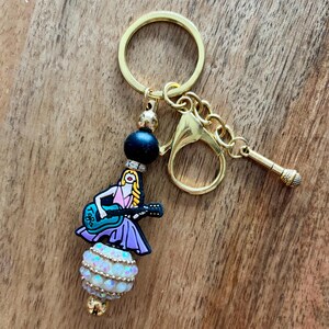 May include: Gold-tone keychain with a charm featuring a cartoon woman playing a blue guitar. The charm is adorned with a black bead, a white and gold beaded sphere, and a small gold microphone charm. The keychain is on a wooden surface.