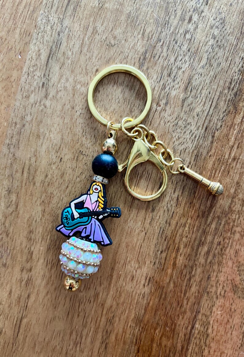 May include: Gold-tone keychain with a charm featuring a cartoon woman playing a blue guitar. The charm is adorned with a black bead, a white and gold beaded sphere, and a small gold microphone charm. The keychain is on a wooden surface.