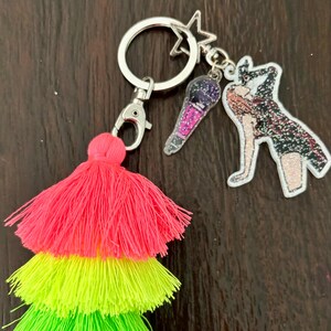May include: A keychain with a silver ring, lobster clasp, and star charm. It features a three-tiered tassel in neon pink, yellow, and green. Additional charms include a microphone and a figure with glitter accents.