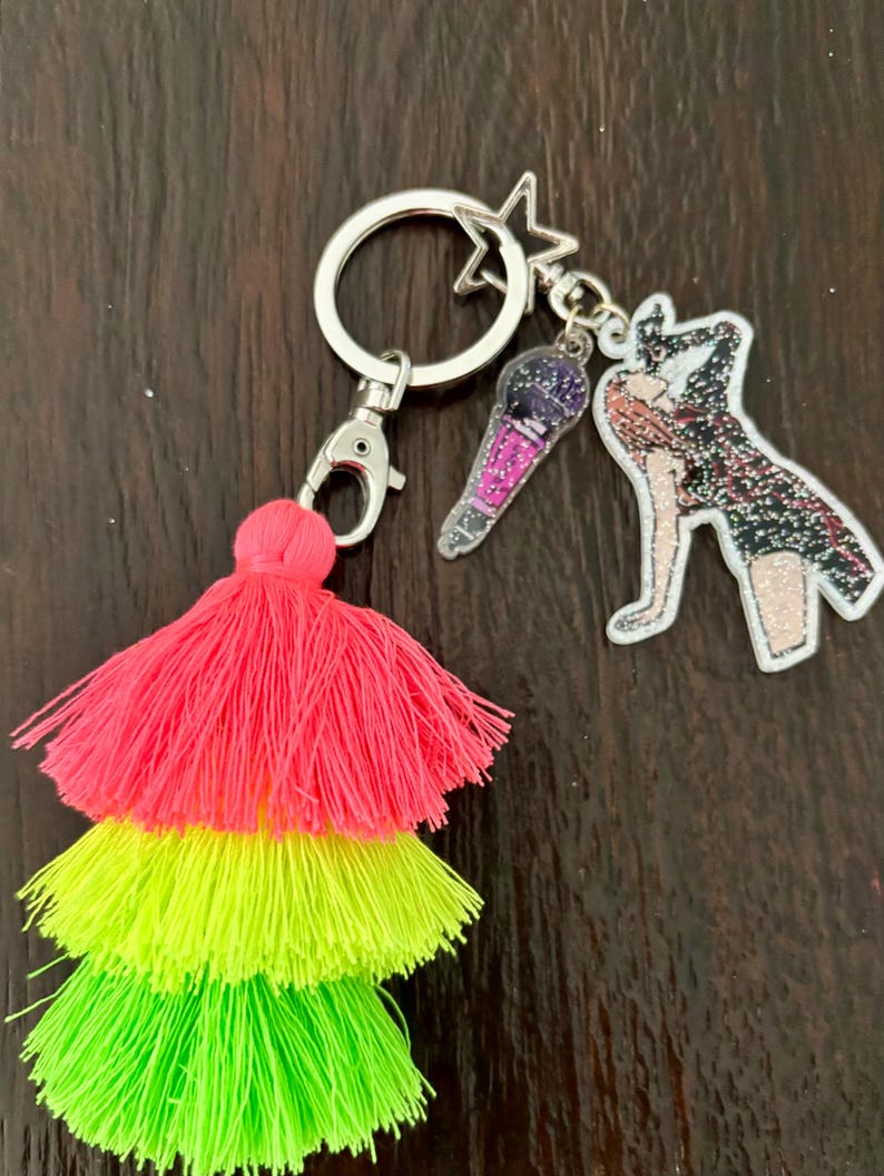 May include: A keychain with a silver ring, lobster clasp, and star charm. It features a three-tiered tassel in neon pink, yellow, and green. Additional charms include a microphone and a figure with glitter accents.