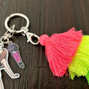 May include: A silver keychain with a singer and microphone charm, and a vibrant pink, yellow, and green tassel. The singer charm is a black and white illustration of a woman singing into a microphone. The microphone charm is pink and purple.