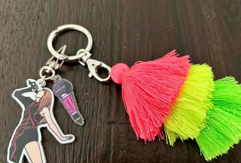 May include: A silver keychain with a singer and microphone charm, and a vibrant pink, yellow, and green tassel. The singer charm is a black and white illustration of a woman singing into a microphone. The microphone charm is pink and purple.