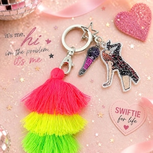 May include: A keychain with a silver ring, star charm, and two dangling charms: a microphone and a figure. A layered tassel in pink, yellow, and green is attached. The text "it's me, hi, I'm the problem, it's me" is visible.