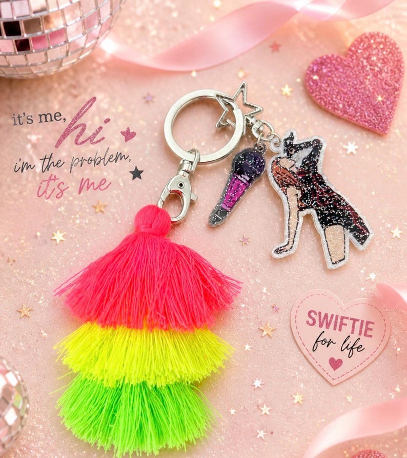 May include: A keychain with a silver ring, star charm, and two dangling charms: a microphone and a figure. A layered tassel in pink, yellow, and green is attached. The text "it's me, hi, I'm the problem, it's me" is visible.