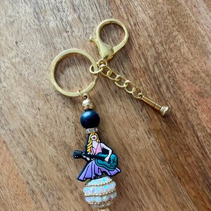 May include: A gold-colored keychain with a charm featuring a cartoon woman playing a guitar. The charm is attached to a beaded design with a black bead, a white and iridescent bead, and a gold-colored clasp and chain. The keychain is on a wooden surface.