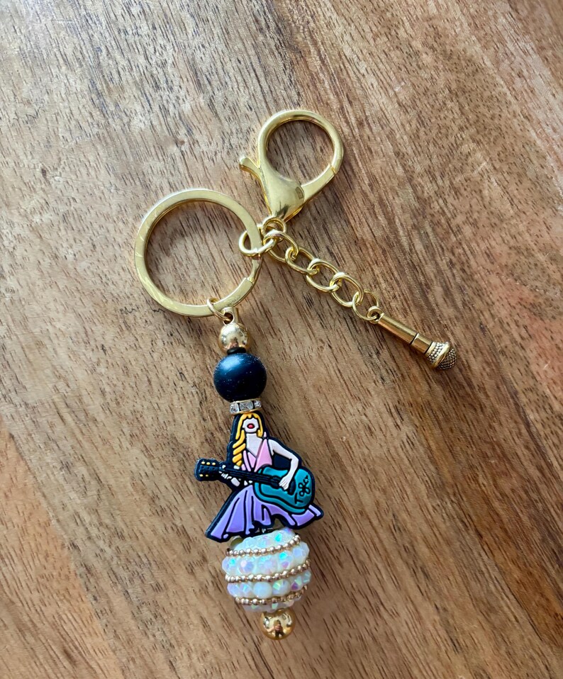 May include: A gold-colored keychain with a charm featuring a cartoon woman playing a guitar. The charm is attached to a beaded design with a black bead, a white and iridescent bead, and a gold-colored clasp and chain. The keychain is on a wooden surface.