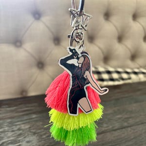 May include: A keychain with a star-shaped clasp and a charm featuring a singer in a black outfit with red accents. The charm is paired with three tiers of neon tassels in pink, yellow, and green.