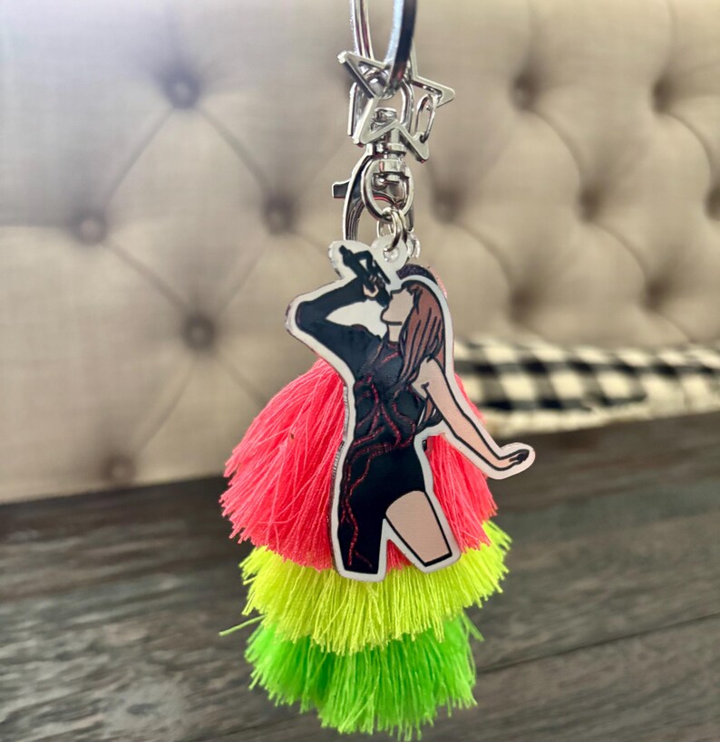 May include: A keychain with a star-shaped clasp and a charm featuring a singer in a black outfit with red accents. The charm is paired with three tiers of neon tassels in pink, yellow, and green.