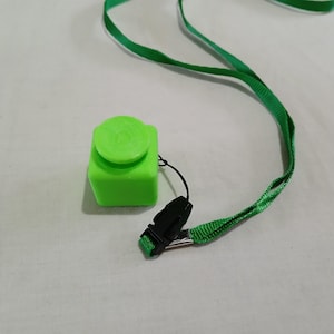 May include: A bright green, square-shaped fidget toy with a round button on top. The toy is attached to a green lanyard with a black clip.