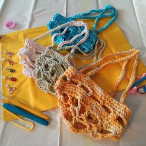 May include: A set of four crocheted bags in different colors, including teal, pink, green, and orange. The bags are made with a mesh pattern and have long handles. The bags are displayed on a yellow surface with crochet hooks and other crafting supplies.