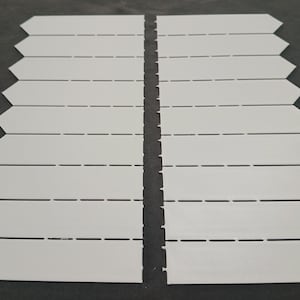 May include: Two sets of white, arrow-shaped, interlocking tiles. Each tile has a rectangular body with a pointed end. Horizontal dashed lines are visible across the tiles. The tiles are arranged on a dark grey surface.