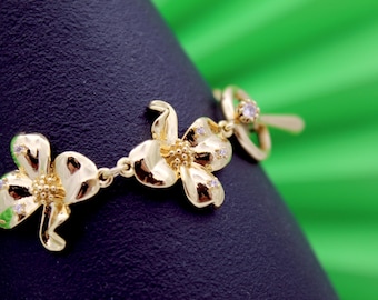 May include: Gold-toned bracelet featuring a floral design. Each flower has three petals and is adorned with small, clear gemstones. The bracelet is displayed against a dark surface with a green background.