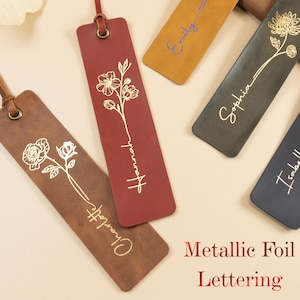 May include: A collection of leather bookmarks in various colors, including brown, burgundy, mustard yellow, dark green, and navy blue. Each bookmark features a floral design and a name in metallic foil lettering. The bookmarks have a leather loop at the top.