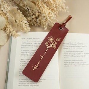 May include: A burgundy leather bookmark with a gold floral design and the name "Hannah" printed vertically. The bookmark has a leather loop and a small leather tassel. The bookmark is resting on an open book.