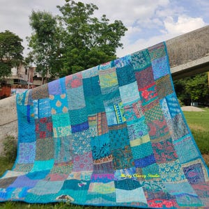 Könnte beinhalten: Ein Patchwork-Quilt in Blau-, Türkis- und Lilatönen mit einer Vielzahl von gemusterten Stoffquadraten. Der Quilt ist über einer Betonkonstruktion drapiert, mit einem Hintergrund aus grünen Bäumen und einem bewölkten Himmel. Der Quilt hat einen blauen Rand.