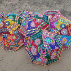 Könnte beinhalten: Eine Ansammlung farbenfroher, dekorativer Regenschirme mit aufwendigen Mustern. Die Schirme weisen eine Vielzahl von Farben auf, darunter Blau, Rot, Gelb und Grün, mit Spiegelakzenten und Fransenrändern. Sie sind auf einer sandigen Oberfläche angeordnet.