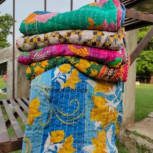Könnte beinhalten: Ein Stapel aus fünf gefalteten Kantha-Steppdecken mit verschiedenen Mustern, darunter Grün mit rosa Blumen, ein schwarz-weißes Karomuster, Magenta mit gelben Blättern, Grün mit gelben Blumen und Blau mit gelben Lilien, gekennzeichnet mit Missyclassystudio.