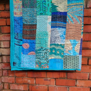 May include: A patchwork quilt made of various blue and brown fabrics with different patterns. The quilt is hanging on a brick wall. The quilt is made of many different pieces of fabric sewn together.