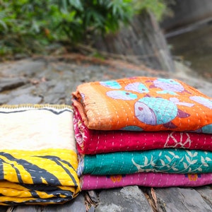 May include: A stack of four colorful kantha quilts with different patterns. The top quilt has a light orange background with blue fish designs. The second quilt is a deep red with a floral pattern. The third quilt is a teal green with a white floral pattern. The bottom quilt is a light pink with a floral pattern.