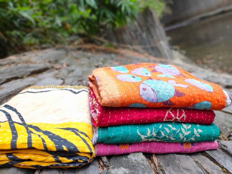 May include: A stack of four colorful kantha quilts with different patterns. The top quilt has a light orange background with blue fish designs. The second quilt is a deep red with a floral pattern. The third quilt is a teal green with a white floral pattern. The bottom quilt is a light pink with a floral pattern.