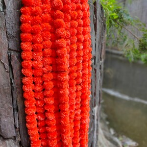 May include: A long garland of bright orange marigold flowers, strung together in a continuous line. The garland is hanging against a stone wall.