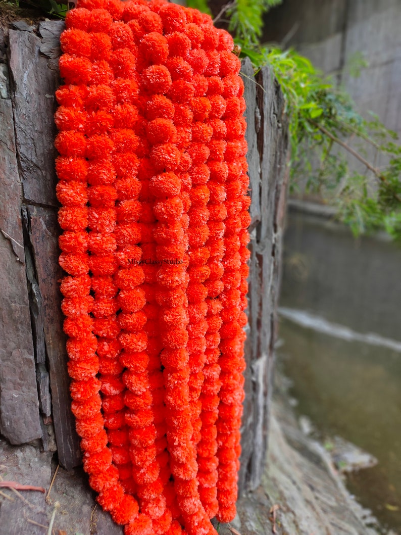 May include: A long garland of bright orange marigold flowers, strung together in a continuous line. The garland is hanging against a stone wall.