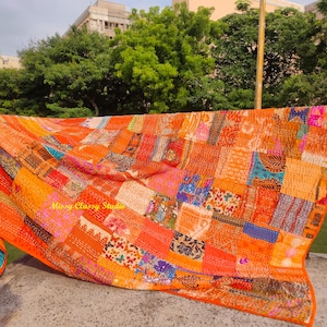 Peut inclure: Une courtepointe patchwork orange vif avec une vari&eacute;t&eacute; de carr&eacute;s de tissu &agrave; motifs. La courtepointe est drap&eacute;e sur une balustrade, mettant en valeur son design complexe et sa palette de couleurs chaudes. Les bords de la courtepointe ont une bordure orange unie.
