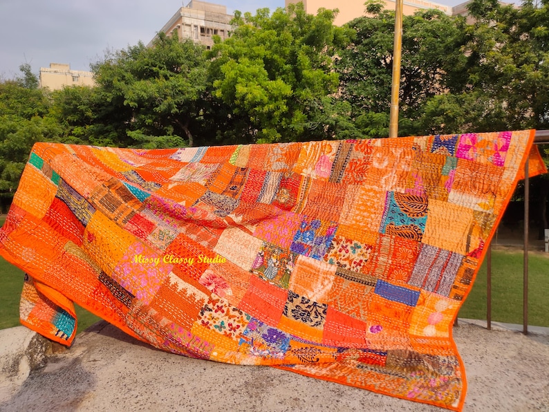 Peut inclure: Une courtepointe patchwork orange vif avec une vari&eacute;t&eacute; de carr&eacute;s de tissu &agrave; motifs. La courtepointe est drap&eacute;e sur une balustrade, mettant en valeur son design complexe et sa palette de couleurs chaudes. Les bords de la courtepointe ont une bordure orange unie.