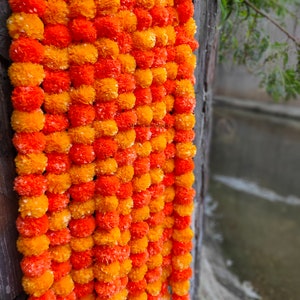May include: A garland of orange and yellow marigold flower pom-poms, hanging against a wooden background.
