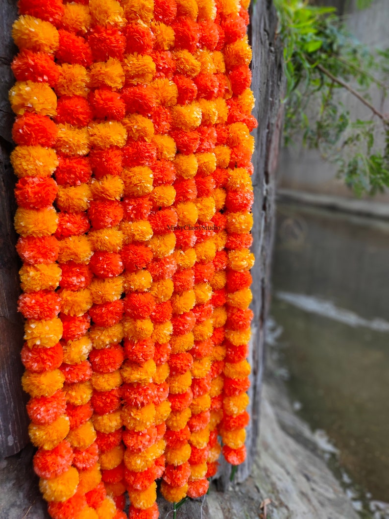 May include: A garland of orange and yellow marigold flower pom-poms, hanging against a wooden background.