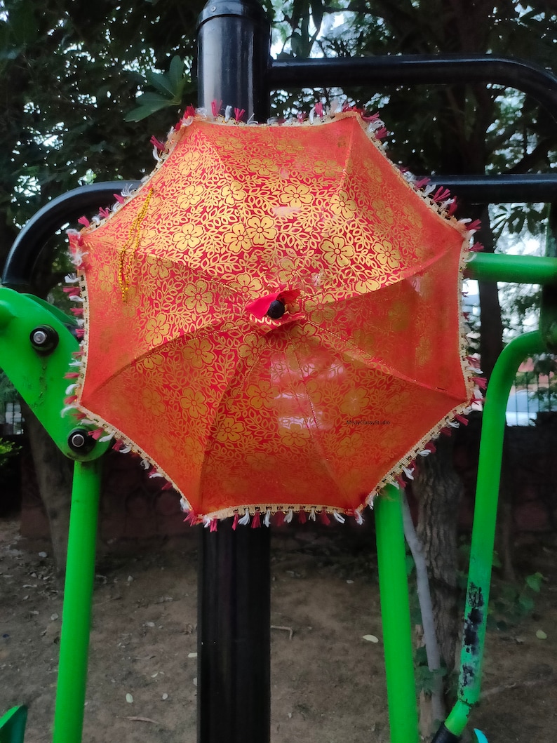 May include: A red and gold patterned umbrella with white fringe hanging from a black pole.