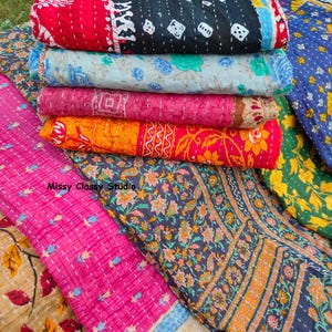 May include: A stack of folded quilts featuring various patterns and colors including red, black, light blue, pink, orange, and dark blue with floral and geometric prints. The text Missy Classy Studio is visible on the fabric. Each quilt has visible white running stitch details throughout the surface.