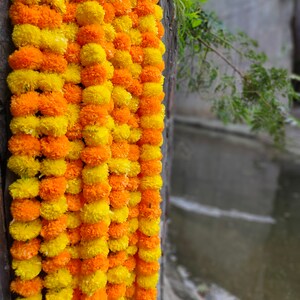 May include: A garland of yellow and orange marigold flowers, strung together in a long strand. The garland is hanging against a wooden surface.