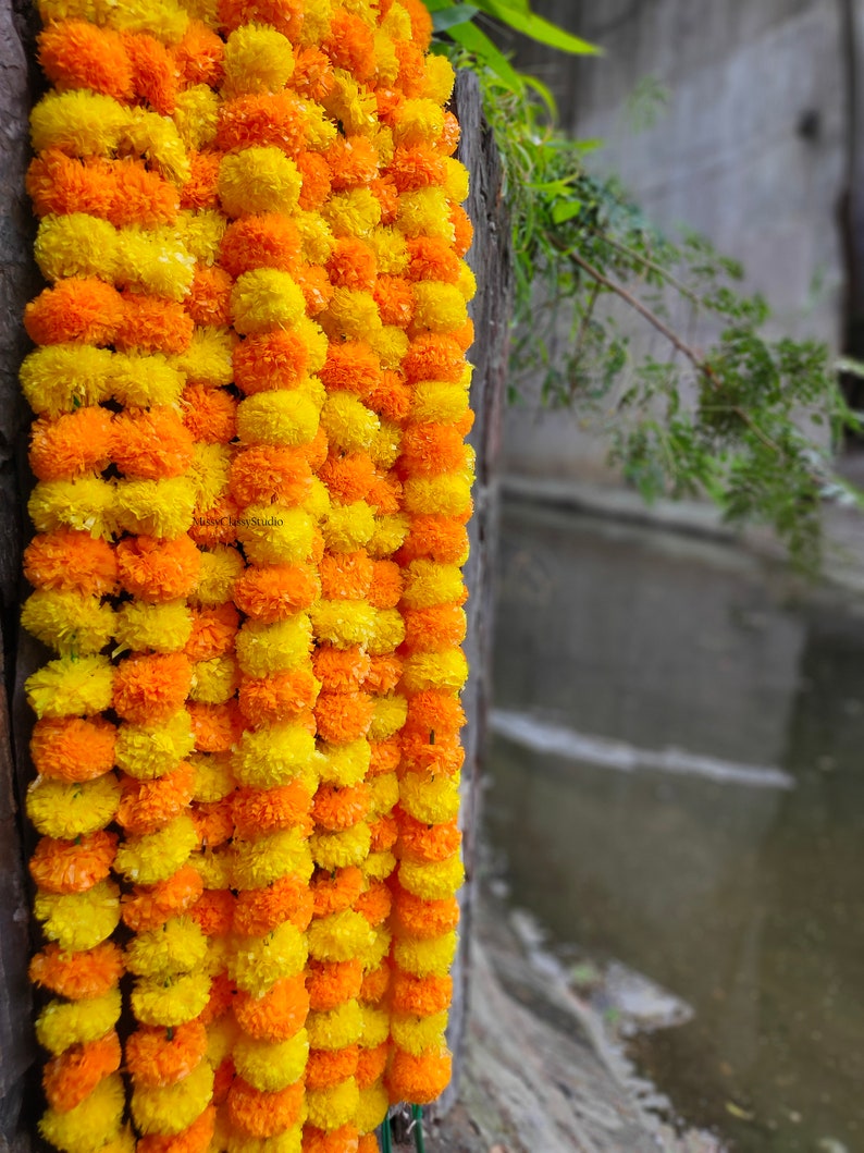 May include: A garland of yellow and orange marigold flowers, strung together in a long strand. The garland is hanging against a wooden surface.