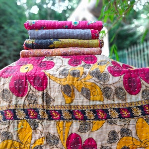 May include: A stack of five kantha quilts in various colors and patterns, including pink, gray, yellow, and orange. The quilts are folded and stacked on top of each other. The top quilt is a pink kantha with a floral pattern. The bottom quilt is a light orange kantha with a paisley pattern.
