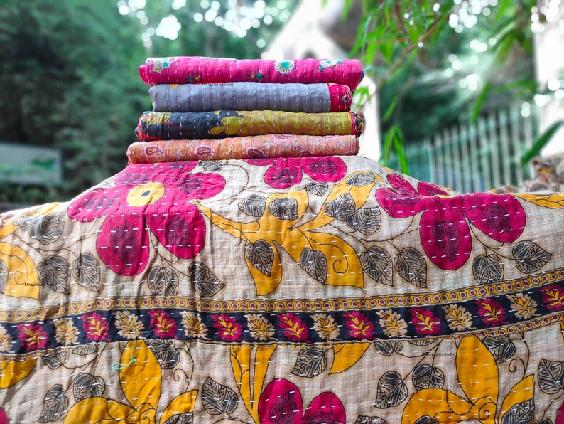 May include: A stack of five kantha quilts in various colors and patterns, including pink, gray, yellow, and orange. The quilts are folded and stacked on top of each other. The top quilt is a pink kantha with a floral pattern. The bottom quilt is a light orange kantha with a paisley pattern.
