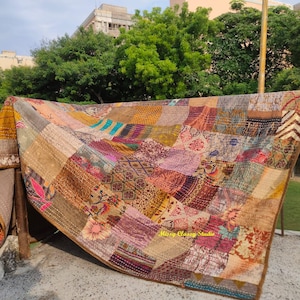 Peut inclure: Une courtepointe patchwork avec divers carr&eacute;s de tissu &agrave; motifs dans des tons de marron, or, rose et bleu. La courtepointe est drap&eacute;e sur une rampe &agrave; l'ext&eacute;rieur, avec une pelouse verte et des arbres en arri&egrave;re-plan. Les bords de la courtepointe sont marron fonc&eacute;.