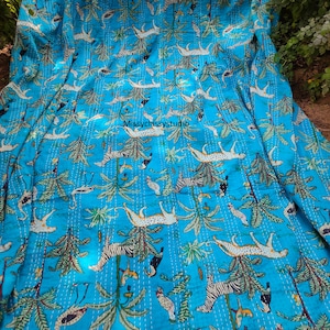 K&ouml;nnte beinhalten: Eine t&uuml;rkisblaue Kantha-Steppdecke mit einem sich wiederholenden Muster aus wei&szlig;en Zebras, Leoparden und V&ouml;geln in einer Dschungelkulisse. Die Steppdecke ist aus Baumwolle gefertigt und hat ein handgesticktes Design. Die Steppdecke ist in der H&auml;lfte gefaltet und liegt auf einer Oberfl&auml;che.