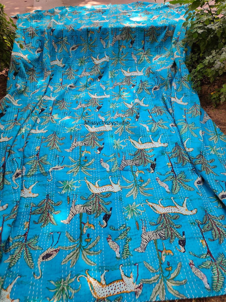 K&ouml;nnte beinhalten: Eine t&uuml;rkisblaue Kantha-Steppdecke mit einem sich wiederholenden Muster aus wei&szlig;en Zebras, Leoparden und V&ouml;geln in einer Dschungelkulisse. Die Steppdecke ist aus Baumwolle gefertigt und hat ein handgesticktes Design. Die Steppdecke ist in der H&auml;lfte gefaltet und liegt auf einer Oberfl&auml;che.