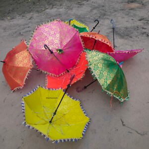 May include: A group of six colorful umbrellas with fringed edges. The umbrellas are made of fabric with gold and silver accents. The umbrellas are arranged in a pile on a sandy surface.