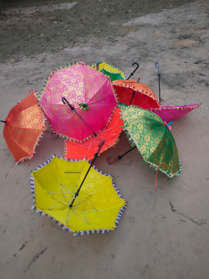 May include: A group of six colorful umbrellas with fringed edges. The umbrellas are made of fabric with gold and silver accents. The umbrellas are arranged in a pile on a sandy surface.