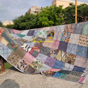 Peut inclure: Une courtepointe patchwork avec une vari&eacute;t&eacute; de tissus dans des tons de bleu, gris, rose et marron. La courtepointe est compos&eacute;e de carr&eacute;s et de rectangles de diff&eacute;rents motifs et textures. La courtepointe est suspendue &agrave; l'ext&eacute;rieur par une journ&eacute;e ensoleill&eacute;e.