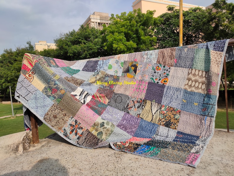 Peut inclure: Une courtepointe patchwork avec une vari&eacute;t&eacute; de tissus dans des tons de bleu, gris, rose et marron. La courtepointe est compos&eacute;e de carr&eacute;s et de rectangles de diff&eacute;rents motifs et textures. La courtepointe est suspendue &agrave; l'ext&eacute;rieur par une journ&eacute;e ensoleill&eacute;e.