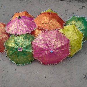 May include: A group of colorful umbrellas with gold and silver accents. The umbrellas are made of fabric and have a fringed trim. They are arranged in a pile on a sandy surface.
