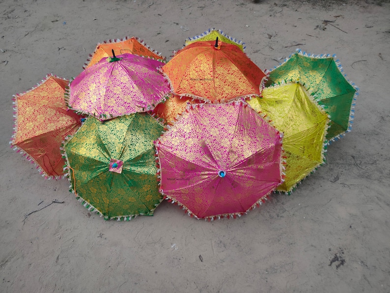 May include: A group of colorful umbrellas with gold and silver accents. The umbrellas are made of fabric and have a fringed trim. They are arranged in a pile on a sandy surface.
