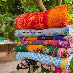 May include: A stack of colorful, quilted blankets. The top blanket is red and orange with floral patterns. Other blankets feature blue, purple, green, and yellow designs. The text "Missy Classy Studio" is visible on one blanket.