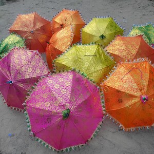 Könnte beinhalten: Eine Sammlung farbenfroher, dekorativer Regenschirme in Orange, Pink, Grün und Gelb. Die Regenschirme haben ein Blumenmuster und sind mit Fransen verziert. Die Regenschirme sind auf einer sandigen Oberfläche angeordnet.